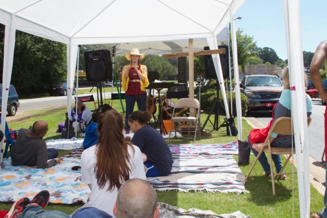 A woman in a wide brim hat under a white tent on a sunny day giving a message about Jesus to people sitting on blankets.