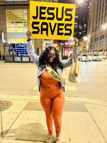 African American woman dressed in a sleek orange jumper with a Birthday Queen sash and tiara, smiling and holding up a yellow and black Jesus Saves sign on the street.