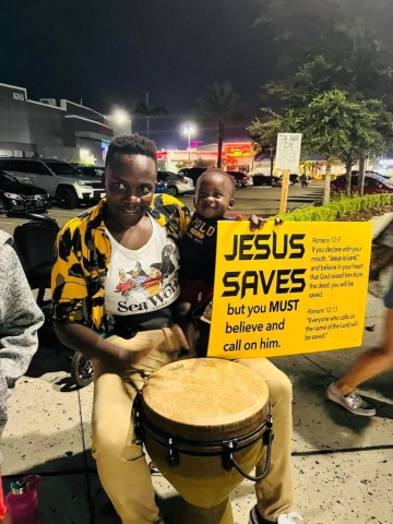 A black man with a baby on his lap playing a bongo on the street at night. He is smiling slightly with a colorful gold and black shirt. He holds a yellow and black sign that says "Jesus Saves but you must believe, call on him, and repent."
