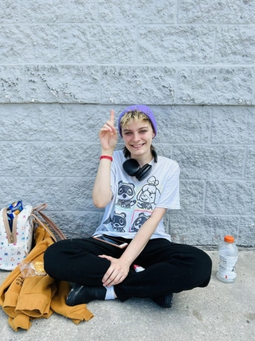 young, thin, hip girl in a purple beanie and big black headphones witting on the street in front of a gray brick wall. She is smiling and pointing up to the sky to show her faith in Jesus.