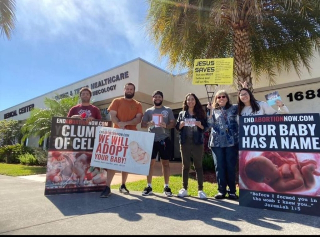 A group of Christians standing on a sidewalk under a palm tree on a sunny day, holding signs about protecting unborn children. One sign says "We will adopt your baby." Another sign says "endabortionnow.com." A lady is holding up a yellow Jesus Saves sign.