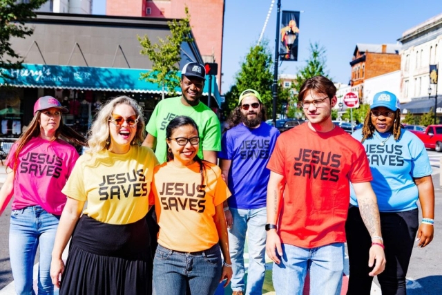various people from different ethnicities walking through a crosswalk wearing brightly colored Jesus Saves shirts in all seven colors of the rainbow.