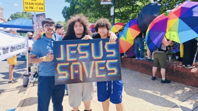 Three young men holding a rainbow sign that says Jesus Saves in a crowd of people with rainbow colored umbrellas.