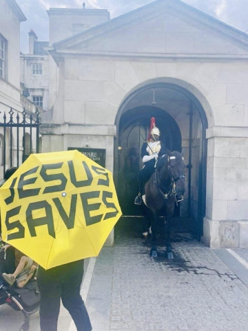 A person holding a yellow Jesus Saves umbrella in front of Buckingham Palace in Britain with the Queen's Guard.