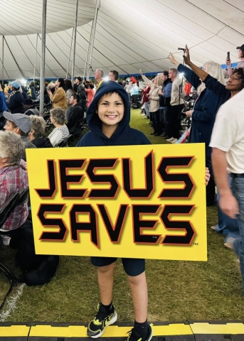 A young boy in a crowd of people worshipping holding up a Jesus Saves yellow sign with red outlines and a drop shadow.
